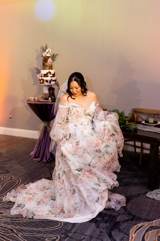 Bride in Floral Off-the-Shoulder Gown with Whimsical Western Flair A bride twirls in a flowing floral off-the-shoulder gown during a western-inspired styled wedding shoot, with a rustic cake table and warm lighting in the background.