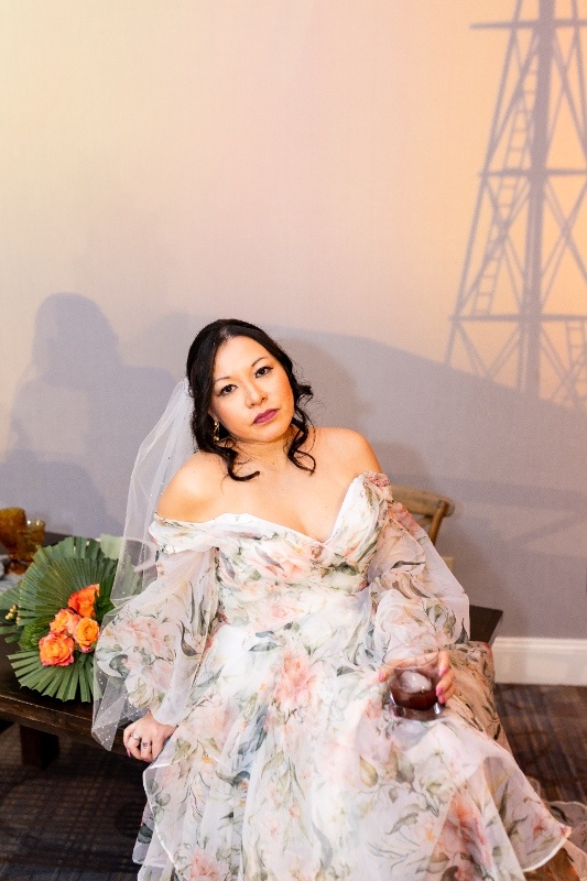 Relaxed Bridal Portrait with Modern Western Vibes A bride in a floral gown and veil sits gracefully, holding a drink and embodying effortless western charm against a rustic Oklahoma!-inspired backdrop.