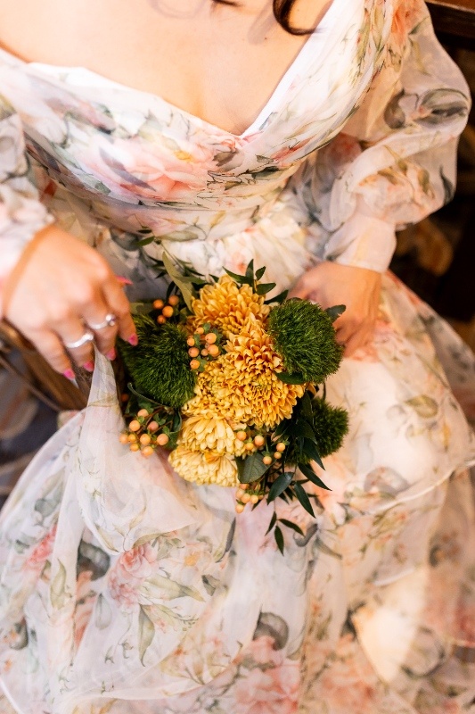 Bride Holding a Rustic Yellow and Green Bouquet Close-up of a bride in a floral off-the-shoulder gown holding a bouquet of yellow mums, green moss balls, and berry accents, capturing the warm rustic tones of this Oklahoma!-inspired wedding shoot.