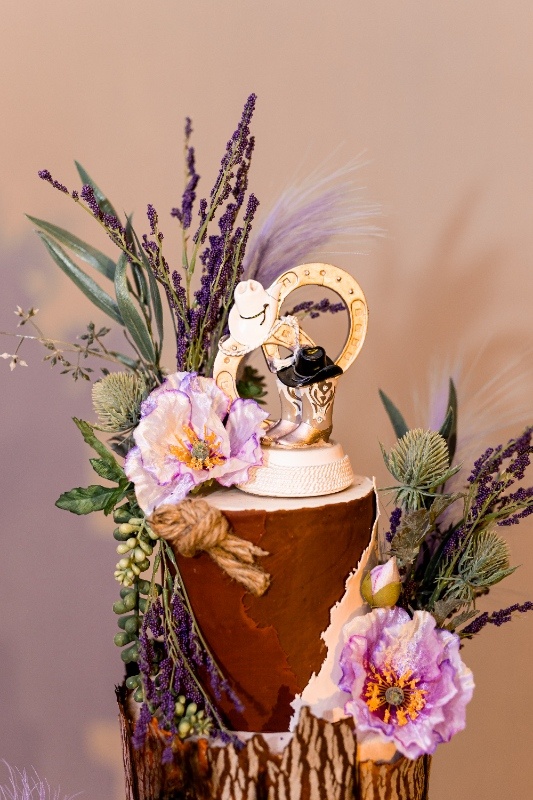 Western-Inspired Cake Topper with Horseshoe and Cowboy Hat Close-up of a rustic cake topper featuring a gold horseshoe, cowboy boots, and hats surrounded by lavender sprigs and soft purple florals, perfect for a western-themed wedding.