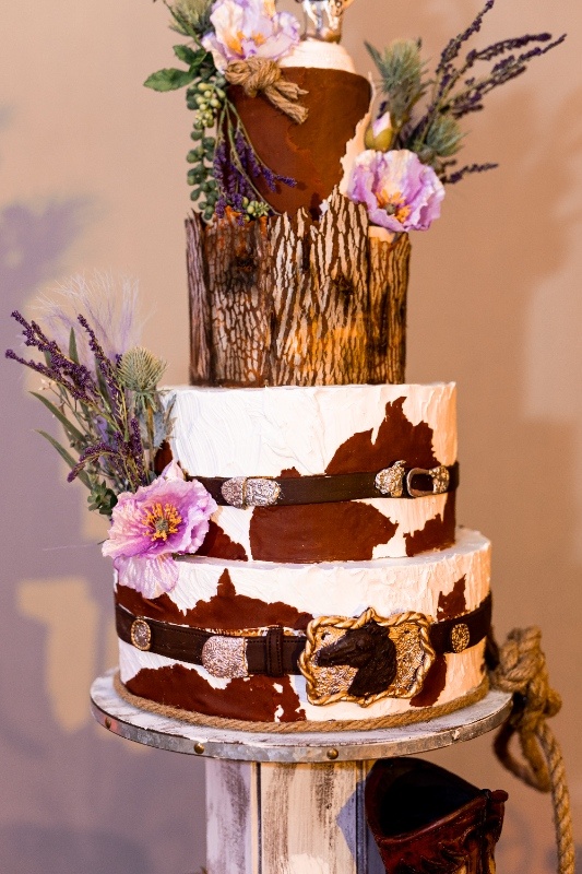 Western Wedding Cake with Leather Belt Details A three-tiered wedding cake featuring cowhide patterns, leather belt accents, and floral embellishments, blending rustic western charm with creative Oklahoma!-inspired design.