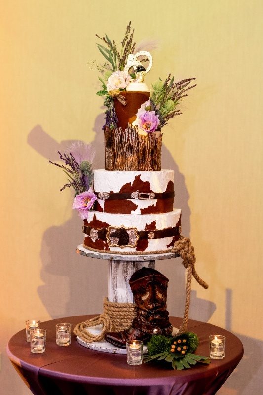 Rustic Western Wedding Cake Display A tall three-tiered wedding cake decorated with cowhide print, leather belts, and purple florals sits atop a wooden table with candles, evoking rustic Oklahoma! wedding inspiration.