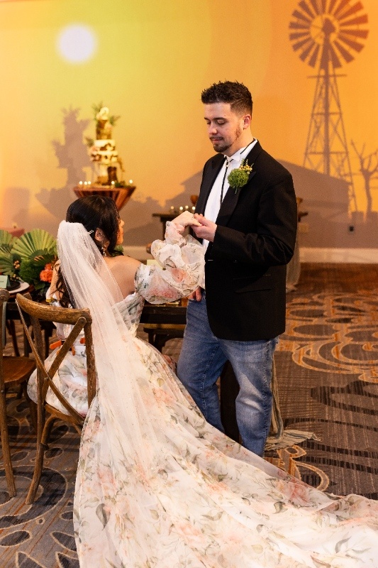 Bride and Groom in Romantic Oklahoma!-Inspired Wedding Scene The bride in a flowing floral gown and the groom in jeans and a blazer share a sweet moment under warm lighting, surrounded by rustic decor and a windmill backdrop inspired by Oklahoma!