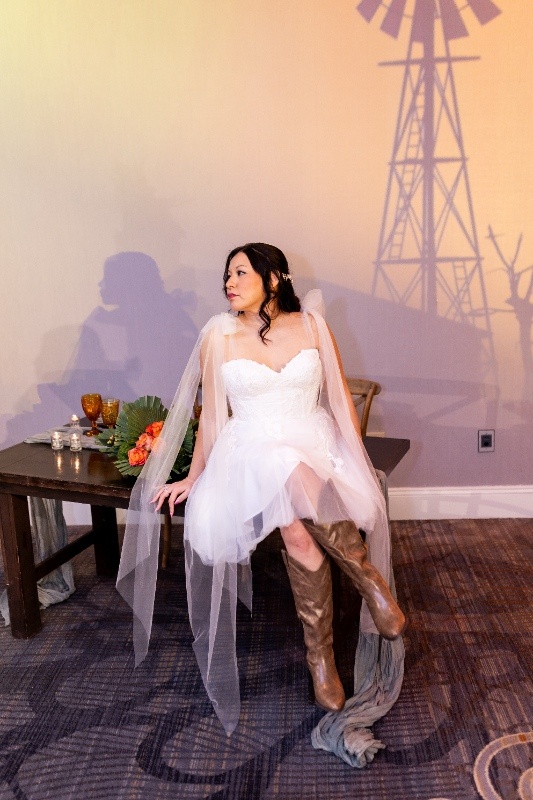 Country-Chic Bridal Portrait with Boots The bride sits gracefully in a short lace gown and tall brown boots against a rustic backdrop, highlighting the fun and flirty side of Oklahoma! wedding inspiration.