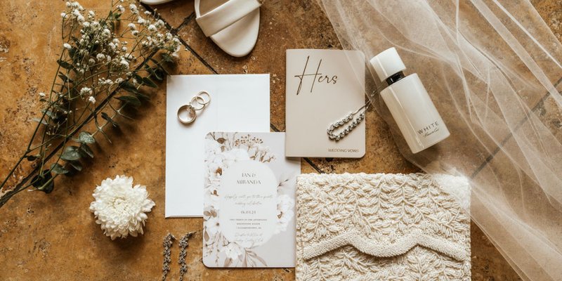 Wedding detail flat lay featuring invitation suite, vow book, rings, bridal jewelry, perfume, veil, and shoes styled with florals.