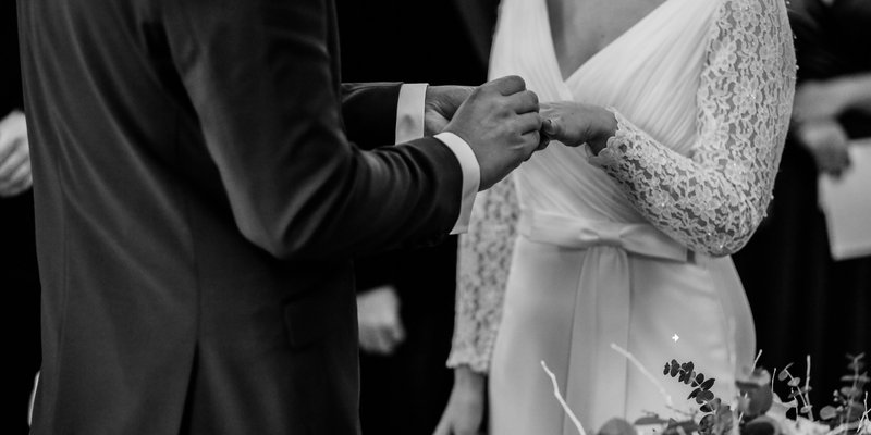 Black and white photo of a couple exchanging rings during an intimate self-uniting wedding ceremony, featuring a bride in a long sleeve lace wedding dress and a close-up of the couple’s hands.