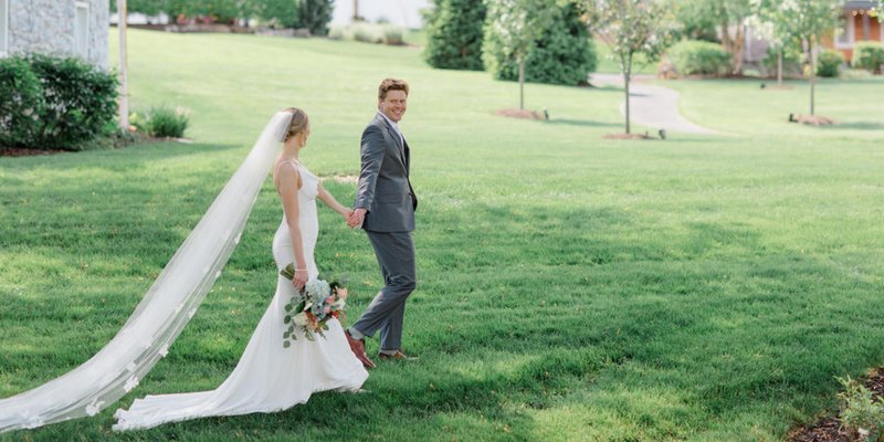 Bride and groom walk hand in hand across the lush green lawn at Historic Ashland, with the bride’s long veil flowing gracefully behind her and the groom smiling back at the camera.