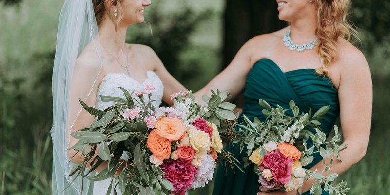 Bride and her maid of honor smiling together while holding colorful bouquets during an outdoor wedding in Central Pennsylvania.