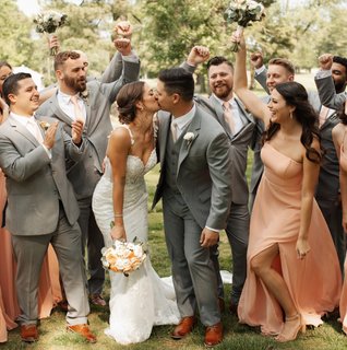 Bride and groom share a kiss surrounded by their bridal party dressed in soft peach bridesmaid gowns and gray suits, celebrating together outdoors at Downingtown Country Club.