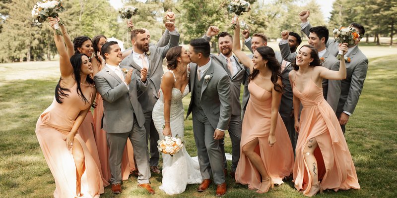 Bride and groom share a kiss surrounded by their bridal party dressed in soft peach bridesmaid gowns and gray suits, celebrating together outdoors at Downingtown Country Club.