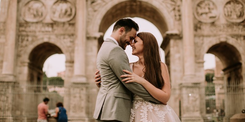 Romantic couple embracing beneath historic European-style stone arches during an intimate destination wedding portrait session, with soft natural light and warm neutral tones.