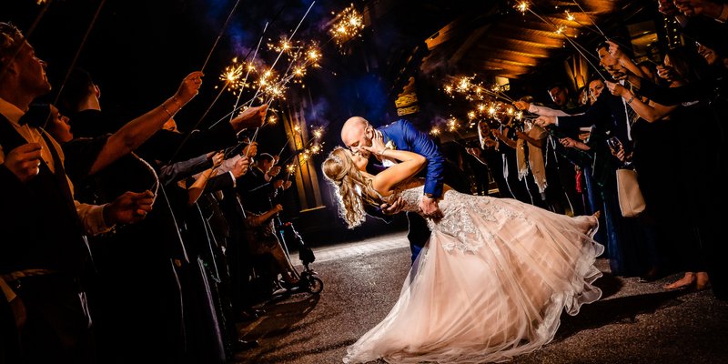 Groom in a blue suit dipping and kissing the bride in the middle of aisle lined with wedding guests holding sparklers