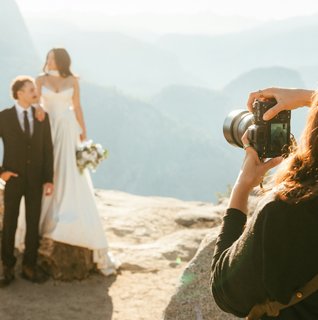 A wedding photographer photographing a bride and groom during an intimate outdoor elopement on a rocky overlook with mountains in the background.
