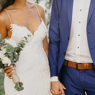 Bride holding white in green florals holding hands of groom in blue suit