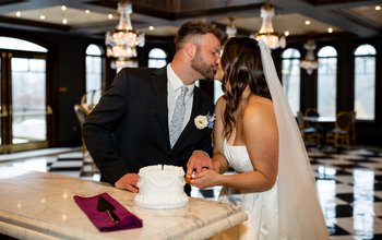 Bride and groom share a kiss while cutting a small white wedding cake on a marble bar inside an elegant reception space with crystal chandeliers, arched windows, and black-and-white checkered floors.
