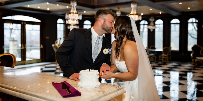Bride and groom share a kiss while cutting a small white wedding cake on a marble bar inside an elegant reception space with crystal chandeliers, arched windows, and black-and-white checkered floors.